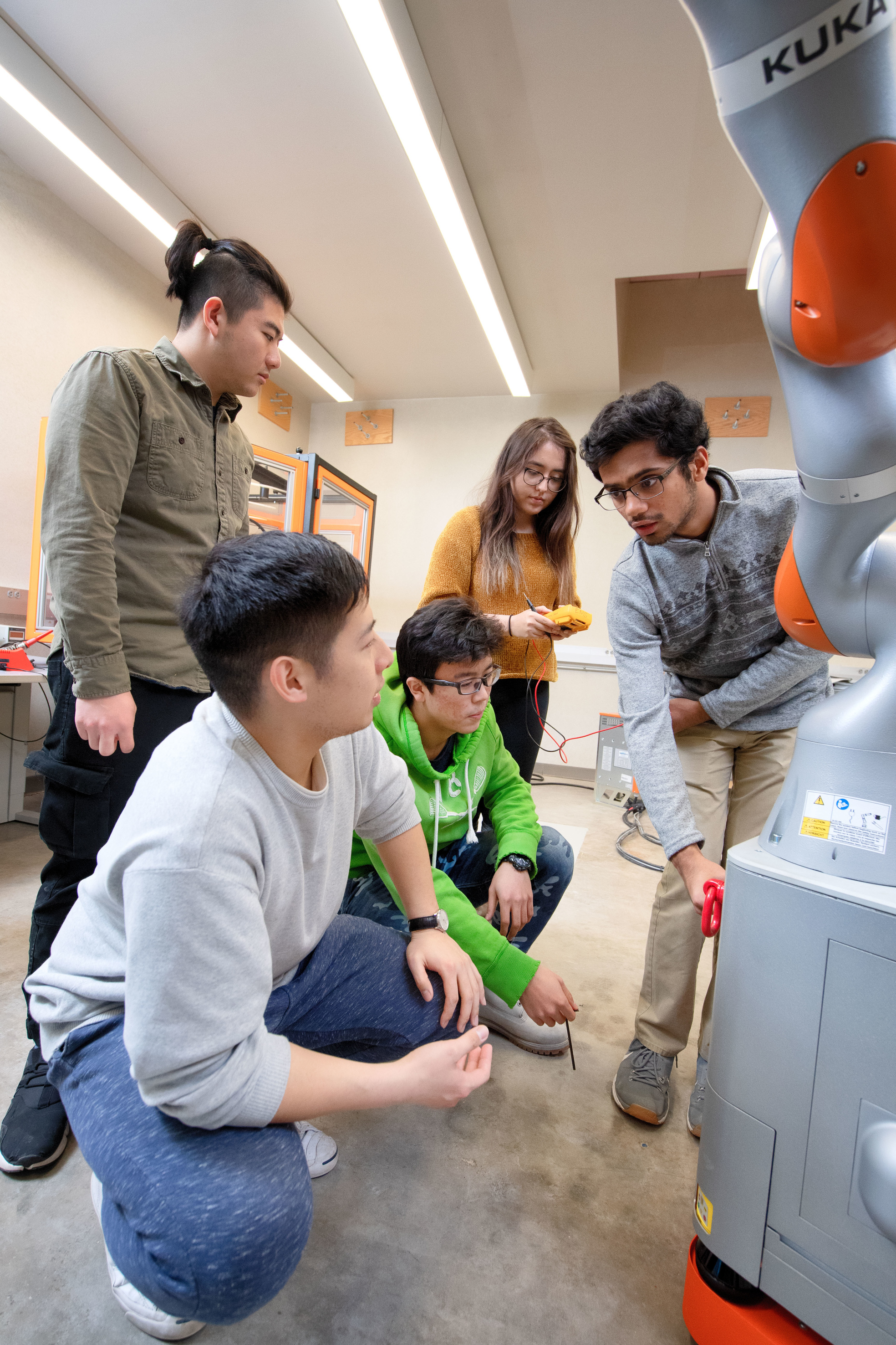 five students crouch and gather around a large gray and orange mechanical arm