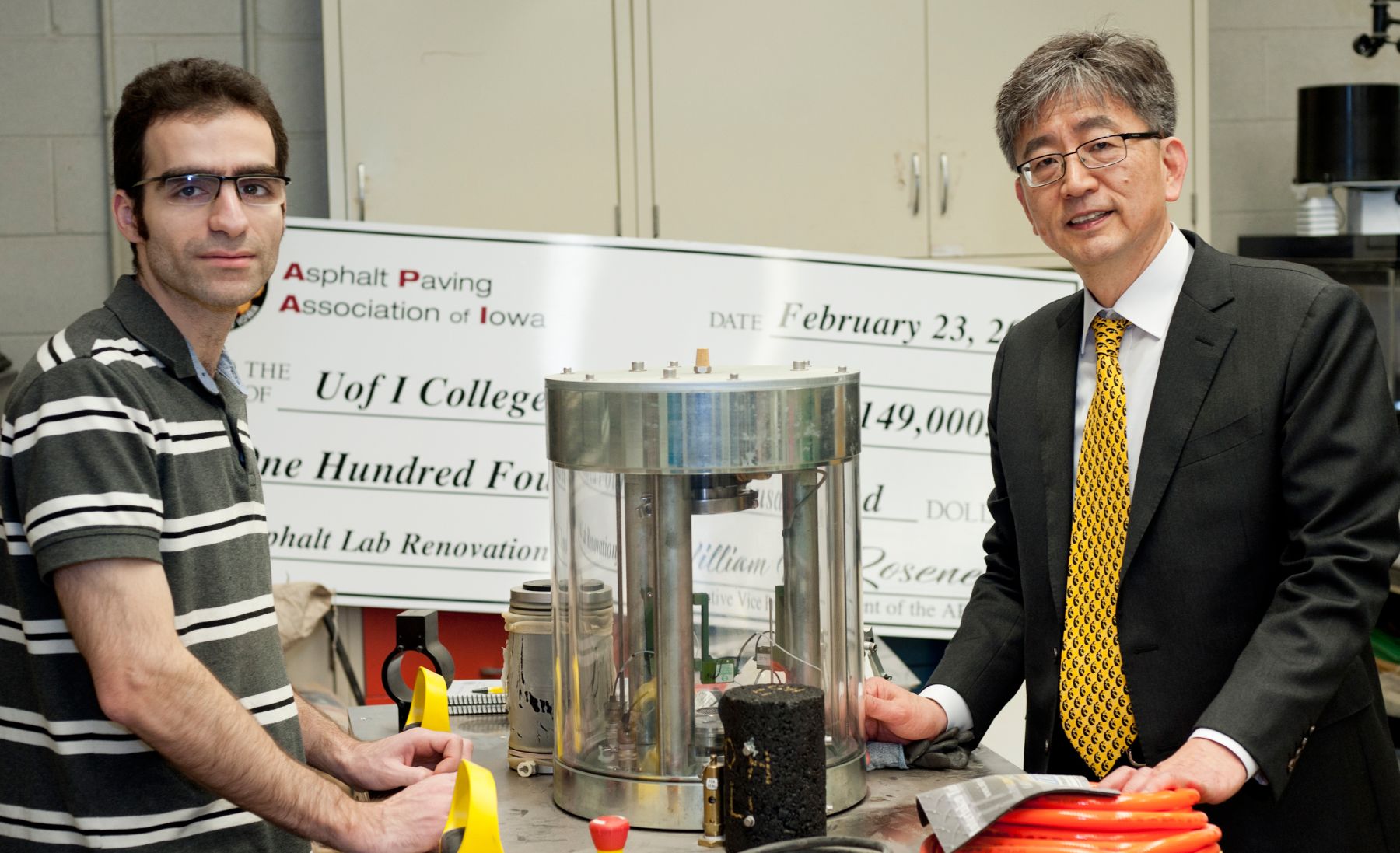 Two people standing at a laboratory workbench with asphalt testing equipment, including a cylindrical testing device and material samples, with a large presentation check from the Asphalt Paving Association of Iowa visible in the background.
