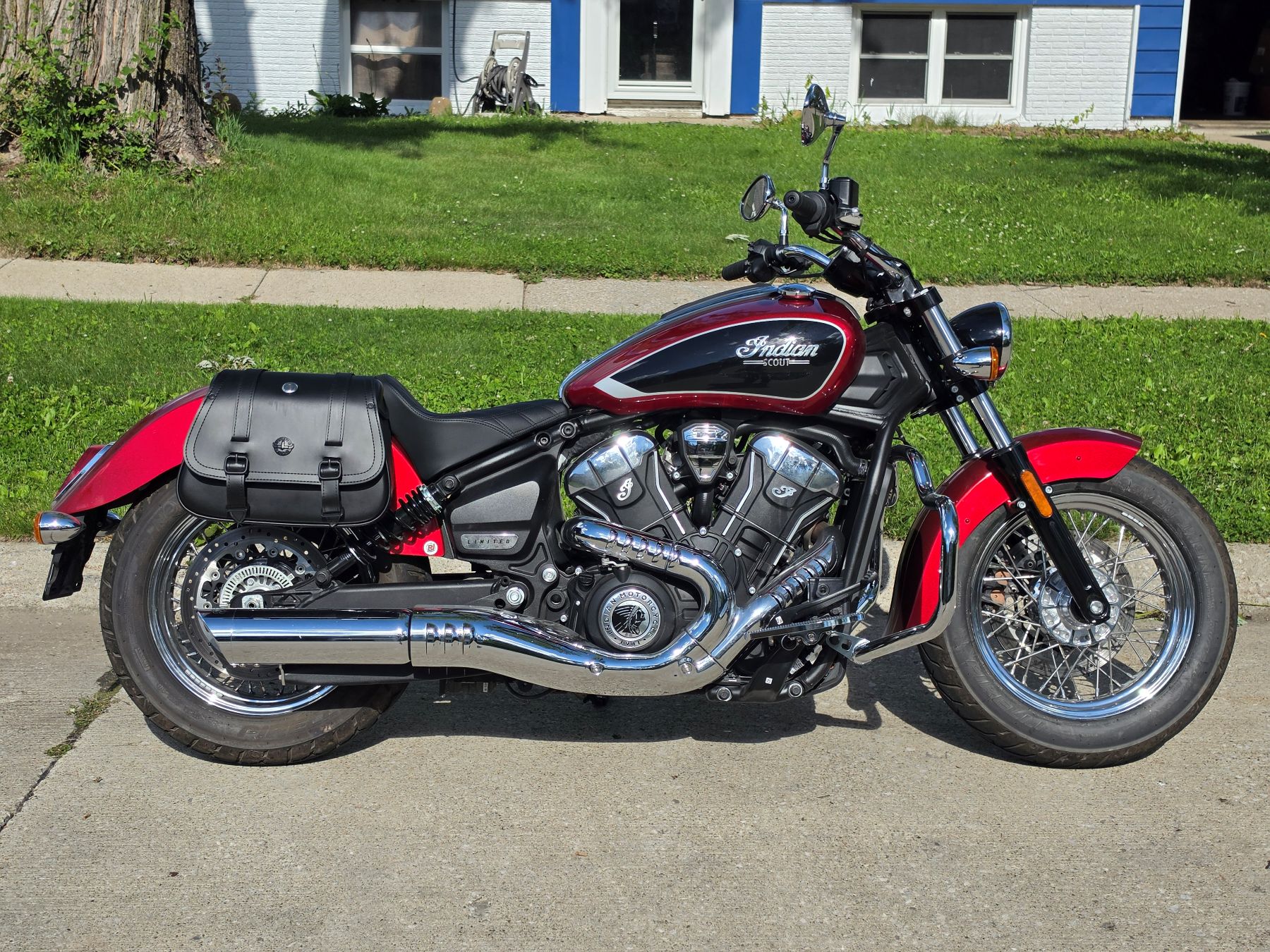 Red and black Indian Scout motorcycle parked on a driveway, viewed from the side with a black leather saddlebag attached.