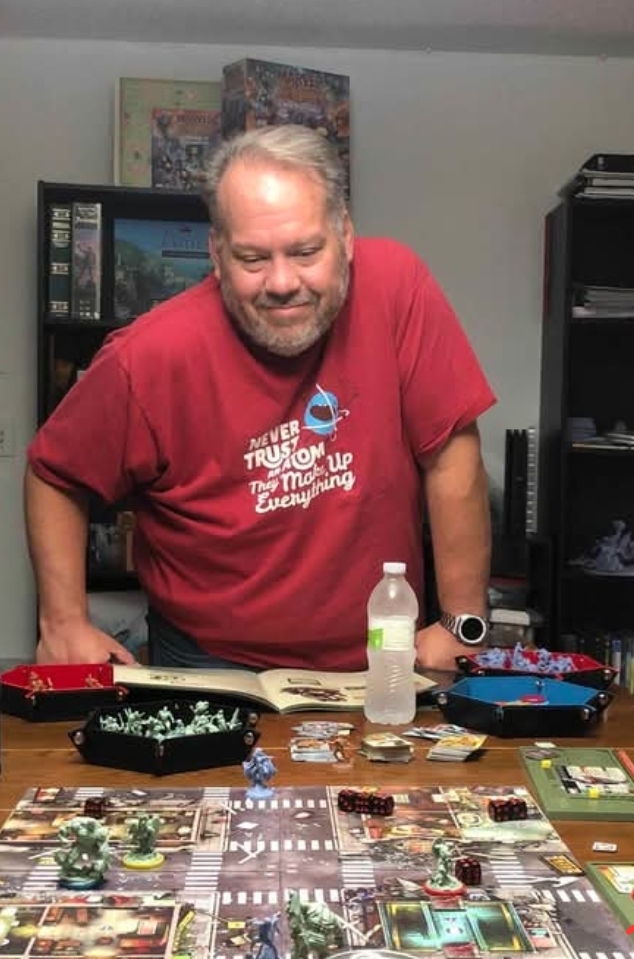 Person standing over a tabletop board game with miniatures, dice, and game cards spread across the playing surface.