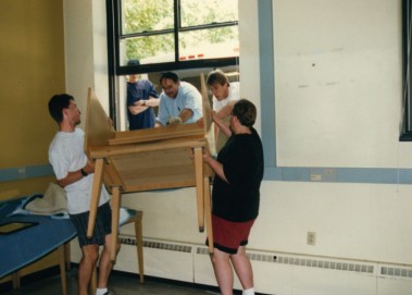 Several people lift a wooden desk through an open window into a room during a library move.