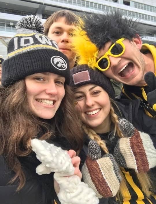 Four people wearing Iowa Hawkeyes winter gear pose for a selfie in the stands of a football stadium.
