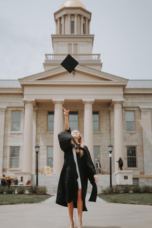 Graduate in cap and gown tosses a graduation cap in front of a classical campus building.