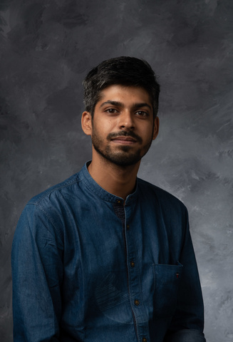 Portrait. Man with dark hair and beard wearing blue button down shirt with studio backdrop.