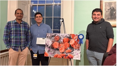 Three men stand by an image of red cells.