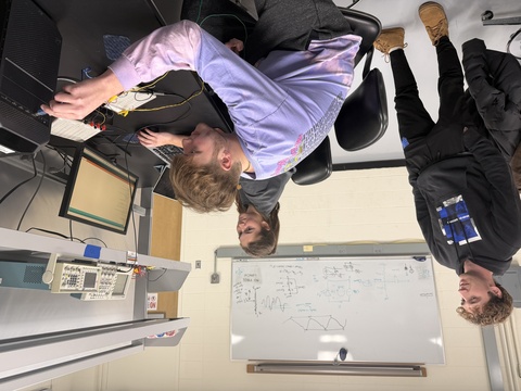 Man in purple shirt reaches for control dials while two other students observe