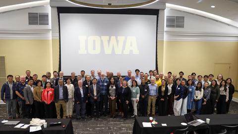 Approximately 75 people stand in front of conference room for photo with "Iowa" displayed on projector screen