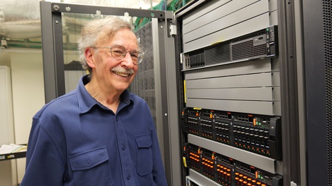 Person standing beside a rack of computer servers and data storage equipment in a server room.