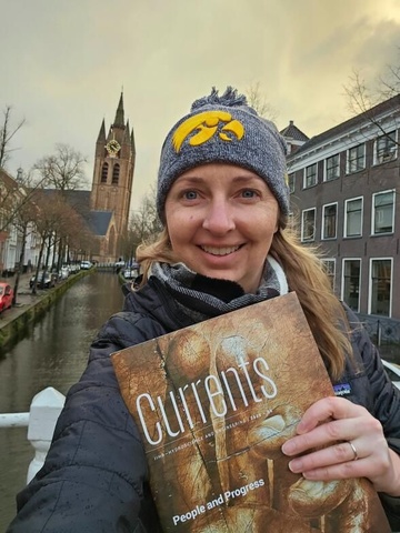 Person wearing an Iowa hat holding a magazine while standing on a canal bridge in a historic city.