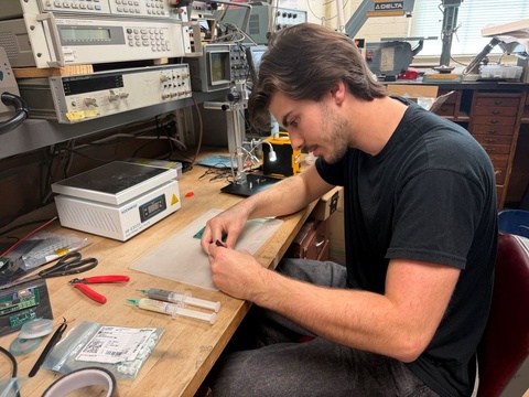 Student assembling a small electronic component at a laboratory workbench surrounded by testing equipment.