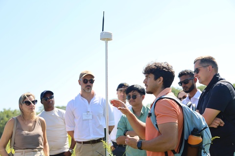 A group of visitors gathers outdoors around a student presenter in a natural field setting. The presenter, wearing a backpack, gestures while explaining a tall environmental monitoring instrument mounted on a pole. Several attendees stand nearby listening, while prairie vegetation and a clear blue sky form the background during the field demonstration.
