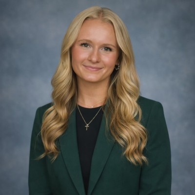 Professional headshot of a person with long wavy blond hair wearing a dark green blazer over a black top and a cross necklace, posed against a gray studio background.
