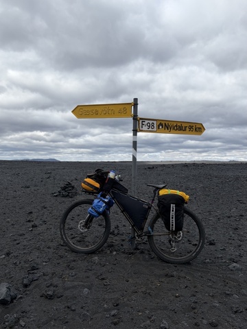 Loaded bikepacking bicycle leaning against a road sign in Iceland’s volcanic highlands, surrounded by dark lava fields beneath a cloudy sky. The sign points to Gæsavötn and Nýidalur.
