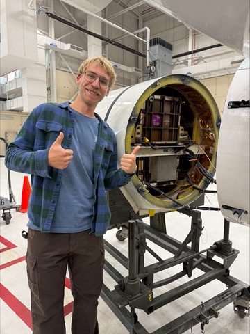 Person standing beside an open aircraft nose compartment, giving two thumbs up while pointing toward a small satellite or avionics payload installed inside a cylindrical housing in a laboratory or hangar setting.
