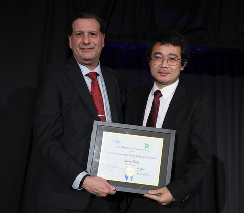 Two people in dark business suits stand on a stage in front of a black curtain, holding a framed certificate. The certificate recognizes the 2026 Du-Co Ceramics Young Professional Award from The American Ceramic Society. One recipient holds the award while posing for a photo during the presentation ceremony.