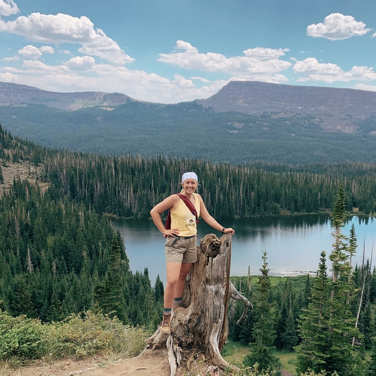 jolee standing on a tree stump in front of a river, a forest, and mountains