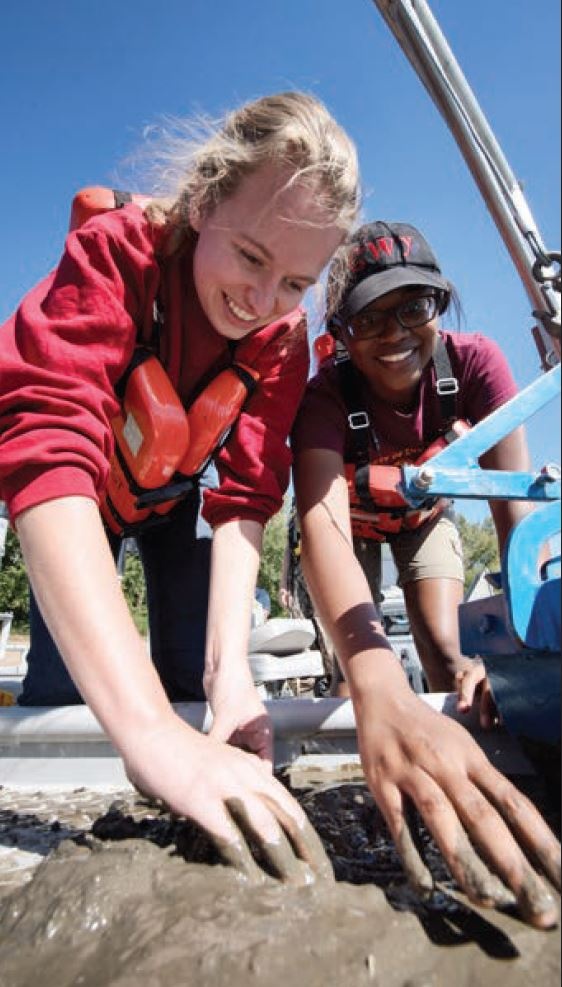 Two students wearing life jackets and working in mud