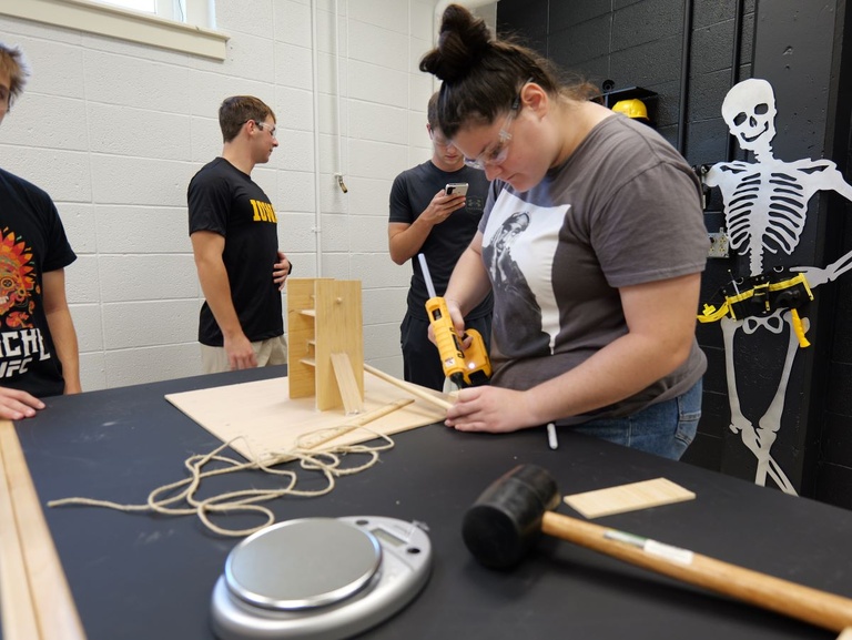 Students in a workshop; one uses a power drill on a wooden project while others observe.