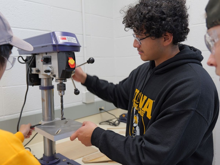 A student operates a drill press while two others observe.
