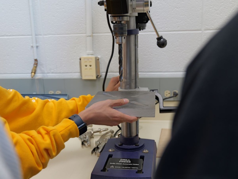 Close-up of hands operating a drill press, securing material for accurate drilling in an educational workshop.