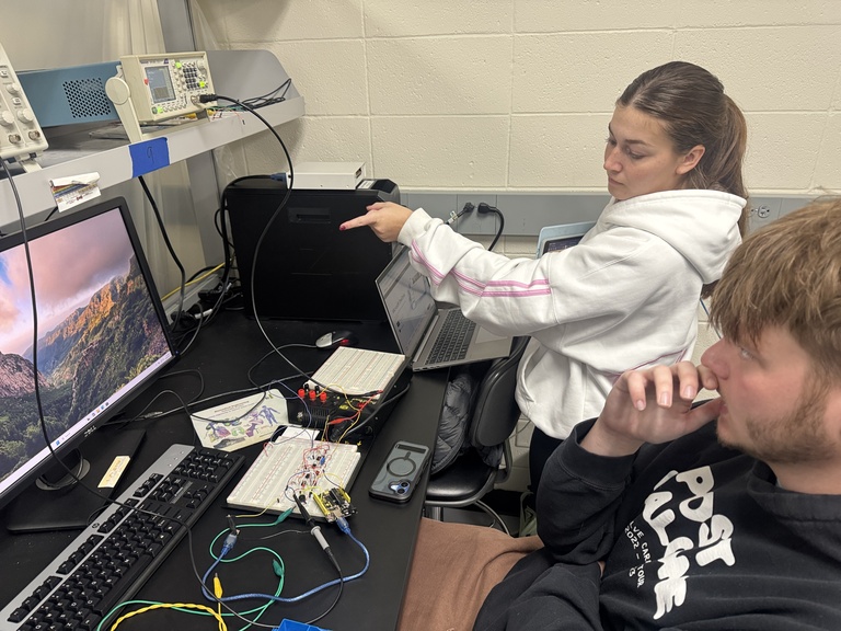 Woman in white shirt points at computer screen while second student looks on
