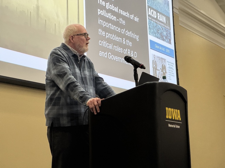 A speaker stands at a podium with an “Iowa” logo, presenting in a lecture hall. A projected slide behind the podium references air pollution, acid rain, and the role of research and government.