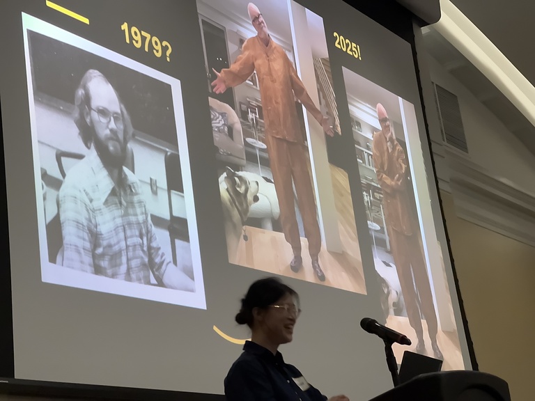 A presenter stands at a podium in front of a projected slide showing three photos labeled “1979?” and “2025!” illustrating changes in appearance over time.