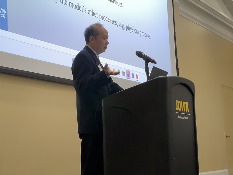 A presenter stands at a podium and gestures while speaking. A projected slide behind the podium displays text related to models and physical processes.