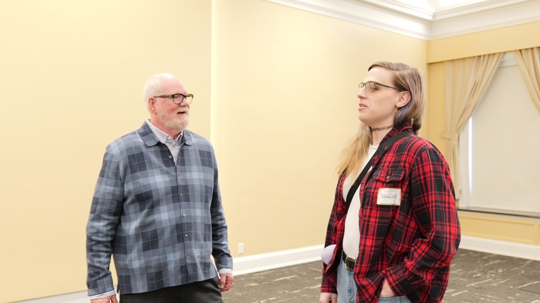 Two attendees stand facing each other and converse in a conference room with pale yellow walls and curtained windows.