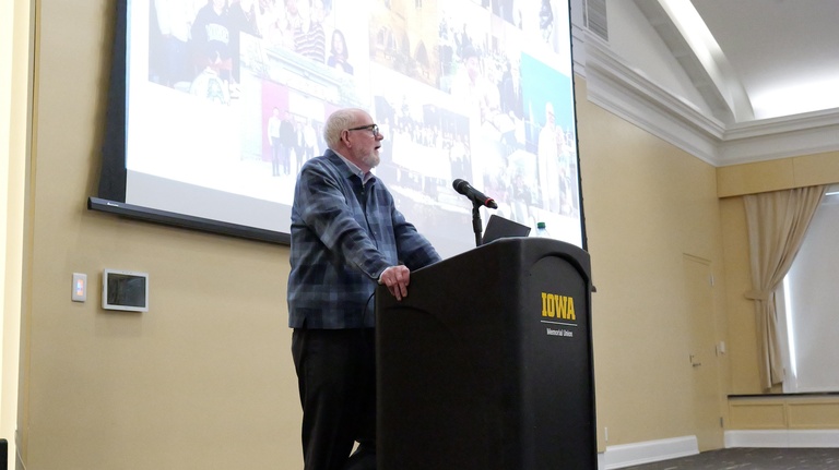 A speaker stands behind an “Iowa” podium in a lecture hall. A projected screen behind the podium shows a collage of multiple photos.