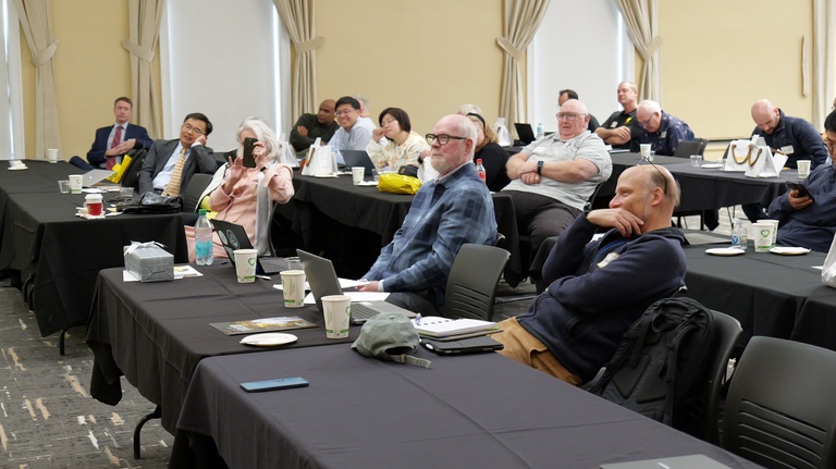An audience sits at rectangular tables in a conference room, listening to a presentation. Laptops, notebooks, cups, and water bottles are visible on the tables.
