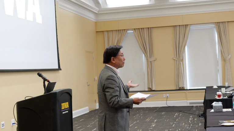 A presenter stands beside a podium while holding papers and gesturing toward the audience in a conference room with tall windows and curtains.