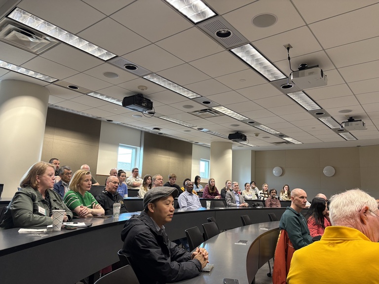 Audience members seated in a curved classroom, facing forward and listening to a panel discussion.