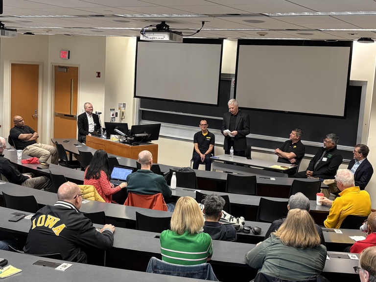 Panel discussion in a tiered classroom, with several speakers seated at the front and one speaker standing, while an audience listens from classroom seating.
