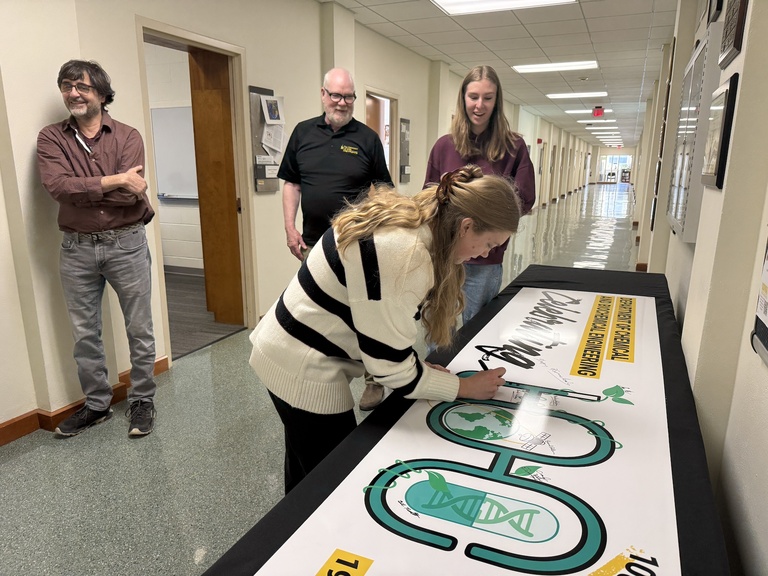 Person signing a large banner laid on a table, featuring prominent lettering and graphic elements, while others observe nearby in a hallway.