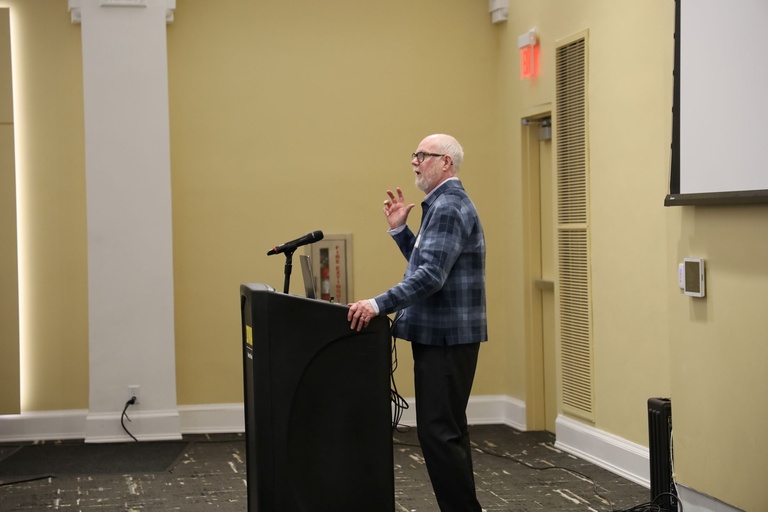 Speaker standing at a black podium with a microphone in a classroom-style hall, gesturing while delivering remarks during the event.