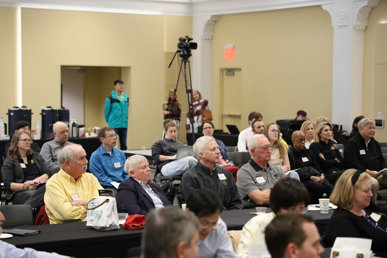 Audience members seated at tables in a lecture hall, listening attentively to a presentation; notebooks, cups, and event materials are visible.
