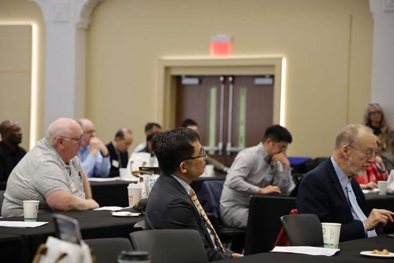 Side view of attendees seated at long tables in a lecture hall, focused on a speaker out of frame during the event program.