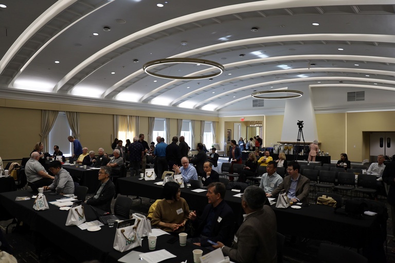Wide view of a large lecture hall filled with people seated at tables and mingling, with tote bags and refreshments visible during the event.
