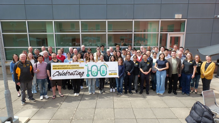 Large group of people standing outdoors in front of a building, posing for a group photo while holding a banner that reads “Department of Chemical and Biochemical Engineering Celebrating 100 Years, University of Iowa, 1926–2026.”