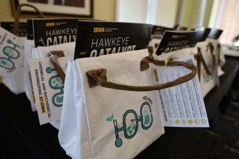 Row of white tote bags on a table, each filled with magazines labeled “Hawkeye Catalyst 2025” and materials from the University of Iowa Department of Chemical and Biochemical Engineering.