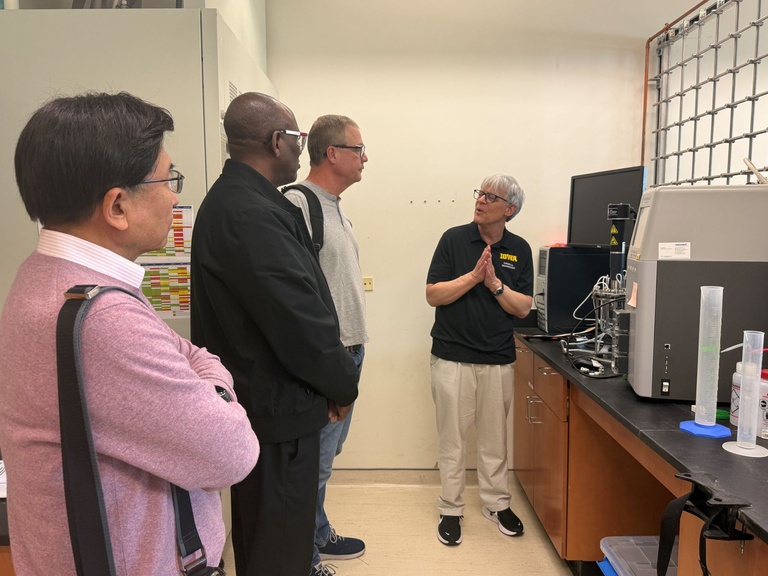 Group of visitors standing in an engineering laboratory while a presenter explains equipment on a lab bench, with instruments, containers, and monitors visible.