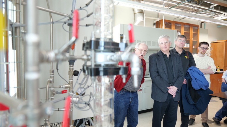 Laboratory tour group standing beside a complex system of pipes, valves, and transparent columns used for engineering experiments.