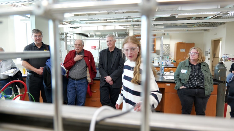 Group of visitors standing in a laboratory while a student demonstrates equipment, with piping and lab benches visible in the foreground.