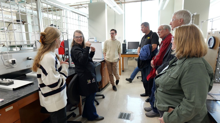 A small group stands in a laboratory as one person gestures while explaining equipment on a lab bench; instruments, tubing, and measuring cylinders are visible.