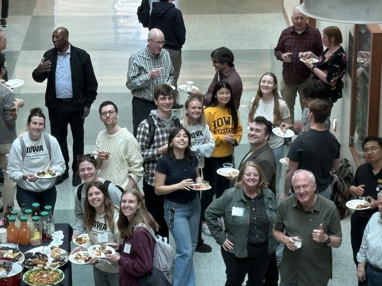 Large group of people standing in an open indoor space, holding food and drinks during a social gathering.