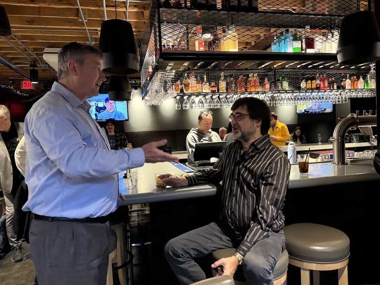 Two people talking at a bar counter, with drinks and bottles displayed on shelves behind them.