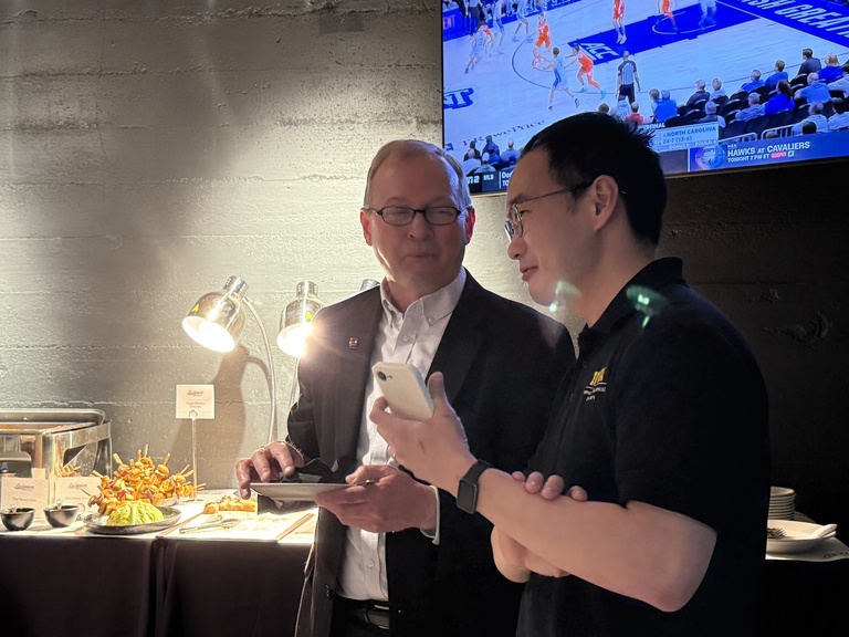 Two people standing near a food station, one holding a phone while plates of food sit under warming lights.