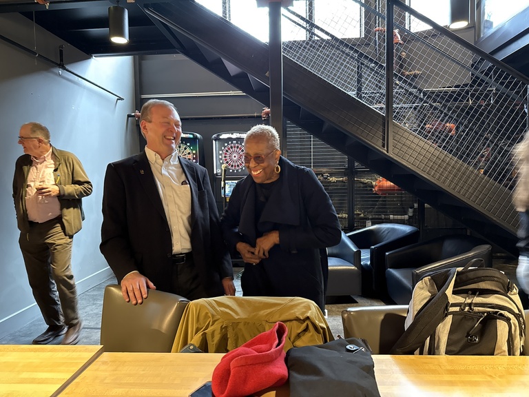 Two people standing together and talking in a lounge area with seating and a dartboard behind them.