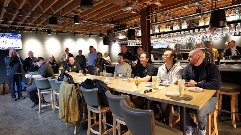 Group of people seated and standing around a long table in a bar-style venue, watching an off-camera speaker.